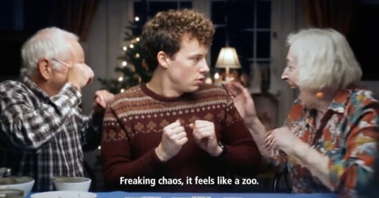 A young man in a patterned sweater sits between two lively older adults at a table, looking startled as they gesture energetically. Subtitles read, "Freaking chaos, it feels like a zoo.