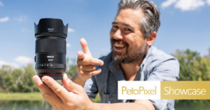 A smiling man outdoors holds up a large camera lens with his left hand and gestures toward it with his right. The logo "PetaPixel Showcase" appears in the lower right corner. Trees and a blue sky are in the background.