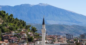A tall white mosque minaret rises among houses and trees on a hillside, with a large green mountain in the background under a clear blue sky.