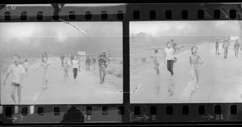Black and white film negatives show children and soldiers running on a road; smoke rises in the background. One child in the foreground appears distressed and unclothed. The scene is tense and chaotic.