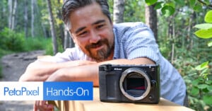 A man with a beard smiles while leaning on a wooden railing in a forest, looking at a Nikon Z camera body placed in front of him. The image includes the text “PetaPixel Hands-On.”.