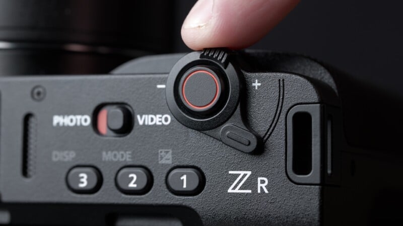 A close-up of a camera's control dial with a red ring being turned by a finger, showing photo and video mode options, and labeled buttons for various settings.