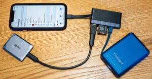 A smartphone connected via adapter hub to various devices including an external hard drive, an SSD, and an SD card, all placed on a wooden surface. The phone screen shows a file explorer app.