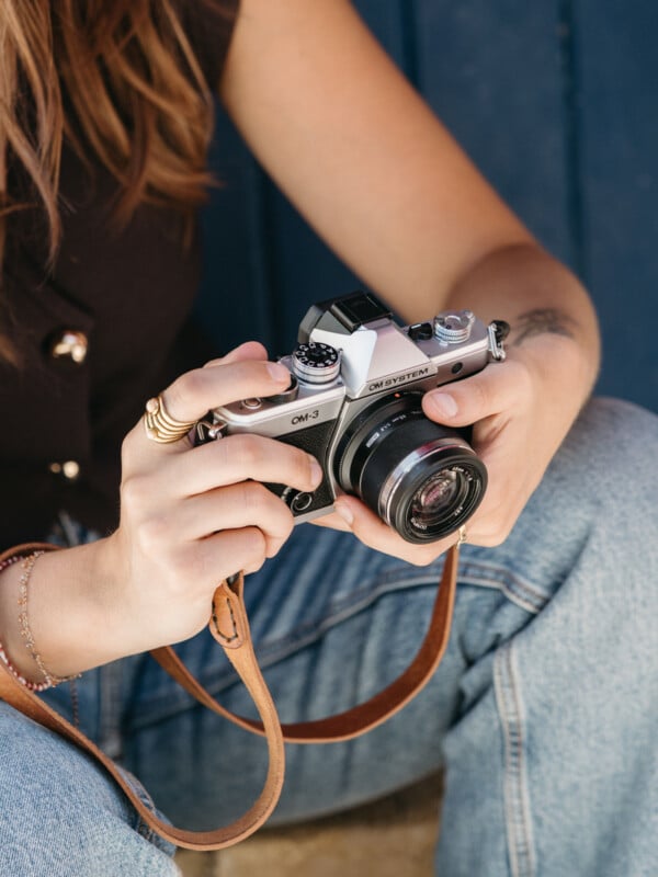 A person wearing jeans holds a vintage Olympus OM-2 film camera with both hands, adjusting its settings. The camera strap hangs down, and the person is seated near a blue background.