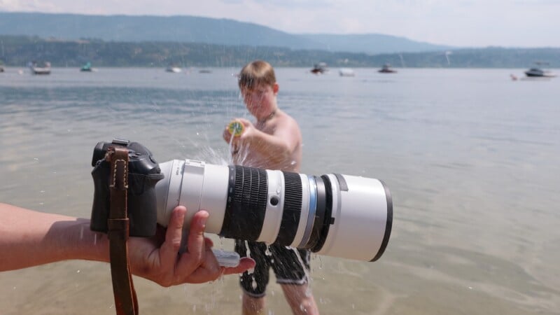 A boy stands in shallow water at a lake, aiming and squirting a water gun toward a large camera with a telephoto lens being held in the foreground. Boats and distant hills are visible under a hazy sky.