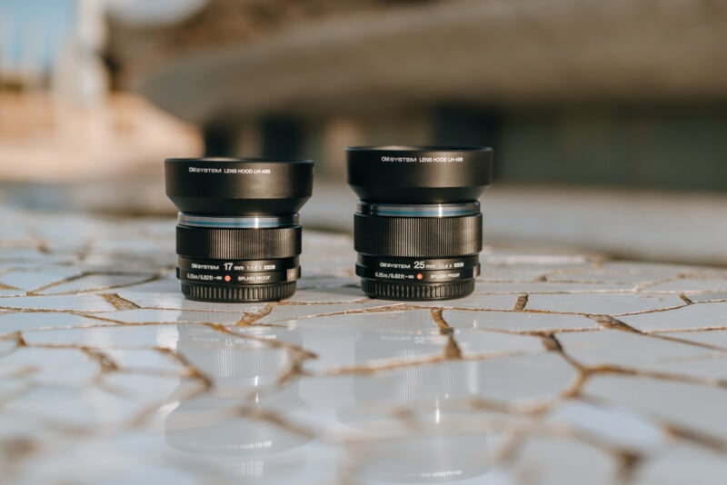 Two Olympus camera lenses stand upright on a tiled white surface with a blurred outdoor background. The lens on the left is labeled 17mm, and the one on the right is labeled 25mm.