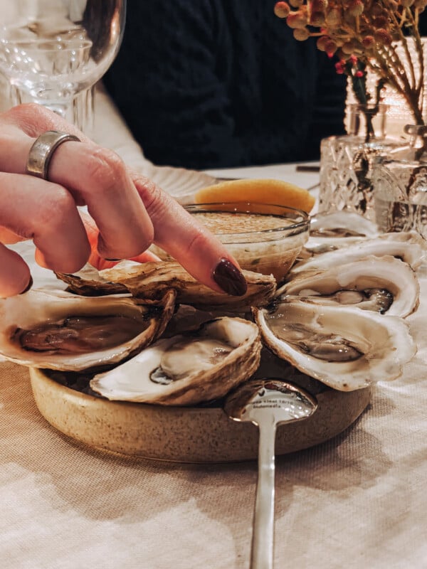 A hand with painted nails and a silver ring lifts an oyster from a plate of oysters on the half shell, with a small dish of sauce, a lemon wedge, and elegant table settings in the background.