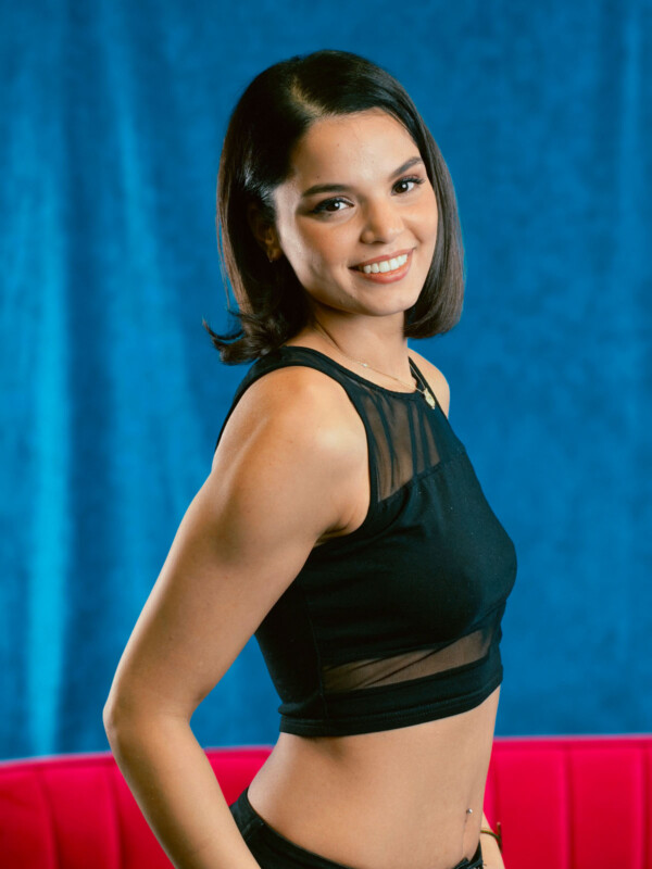 A young woman with straight, shoulder-length dark hair smiles while posing in front of a blue background. She is wearing a sleeveless black crop top and stands slightly turned to the side.