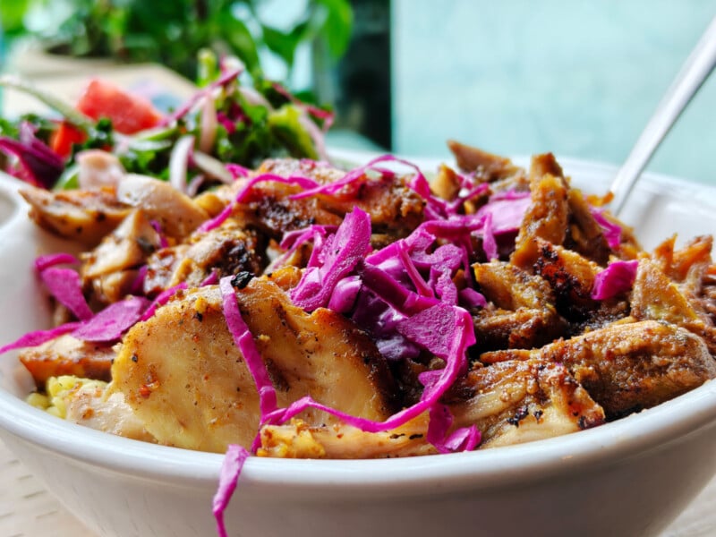 A close-up of a bowl filled with grilled chicken pieces, topped with shredded purple cabbage, and accompanied by a mixed salad in the background. The dish is vibrant and fresh, served in a white bowl.