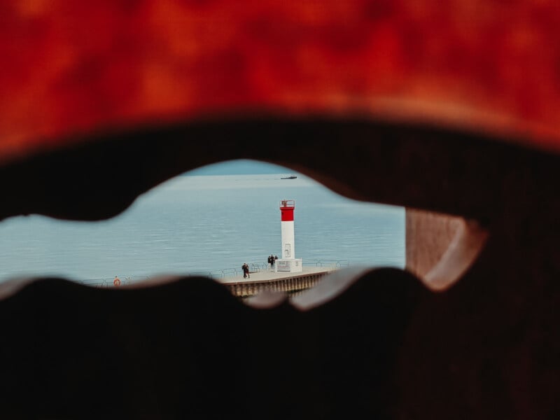 View of a red and white lighthouse by the water, seen through an irregularly shaped opening in a dark foreground object, with a few people walking near the lighthouse and calm water in the background.