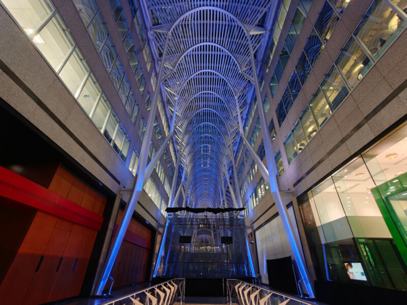 A modern, vaulted interior with tall, white steel arches forming a ribbed ceiling, flanked by glass-walled offices and shops. Blue lighting highlights the arches, creating a futuristic, airy atmosphere.