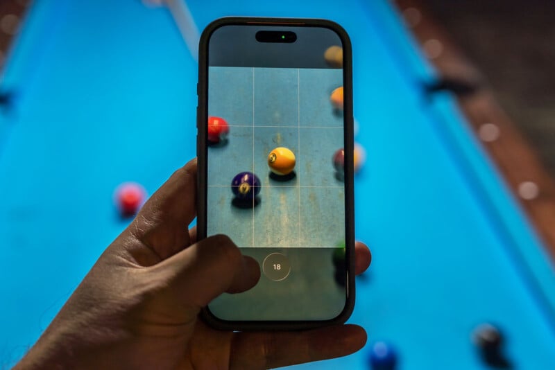 A hand holds a smartphone, taking a close-up photo of billiard balls on a blue pool table. The phone screen displays the balls in focus, while the actual pool table appears blurred in the background.