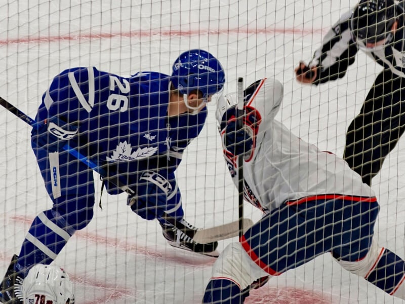 Two hockey players, one in a blue uniform and the other in white, face off for the puck on the ice rink. An official in black and white stripes stands nearby. The scene is viewed through a protective net.