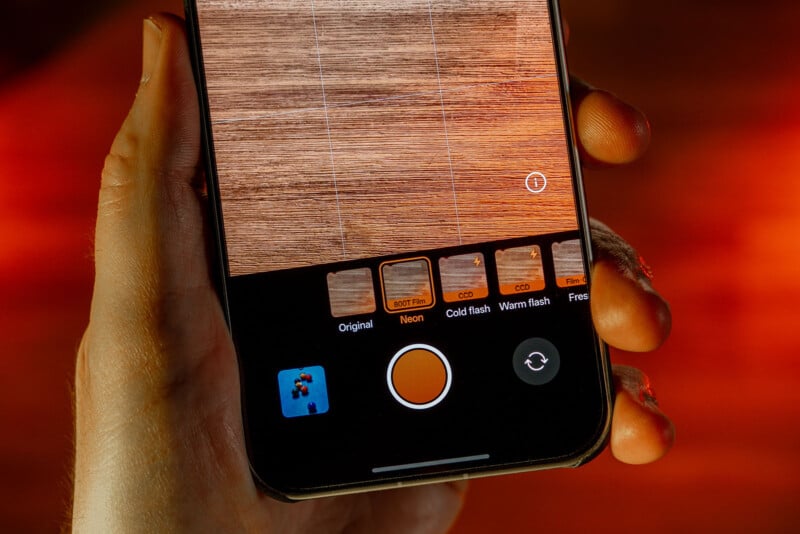A hand holds a smartphone showing a camera app screen with photo filters. The wood surface is visible through the camera, and the interface displays filter options like "Original," "Neon," and flash settings.