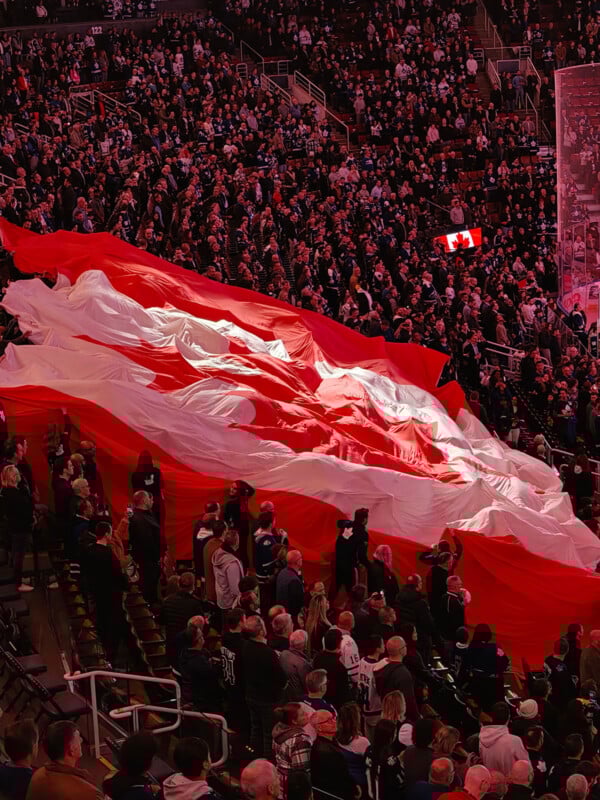 A large crowd in a stadium holds up a giant Canadian flag, with red and white stripes, while many spectators stand and watch. A smaller Canadian flag is illuminated in the background.