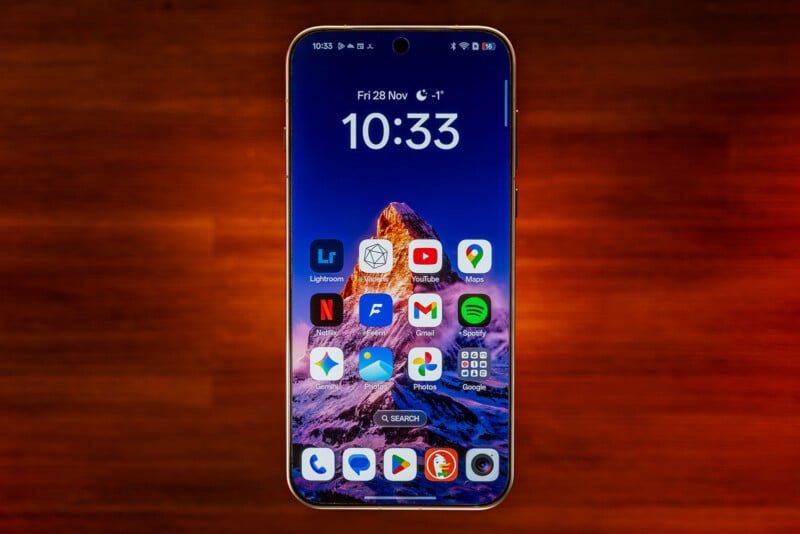 A smartphone with a mountain wallpaper displays the home screen at 10:33 on Friday, November 28. App icons such as Lightroom, YouTube, Maps, Gmail, and Spotify are visible against a brown-orange blurred background.