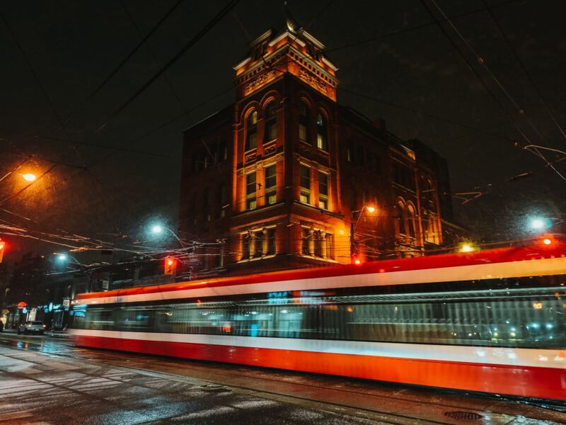 A red and white streetcar moves quickly past a historic brick building at night, with lights reflecting on the wet street and illuminating the scene.