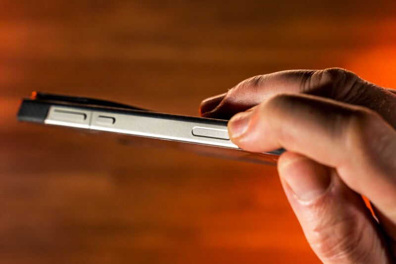 A hand presses the volume button on the side of a slim smartphone, with a blurred warm-toned background.