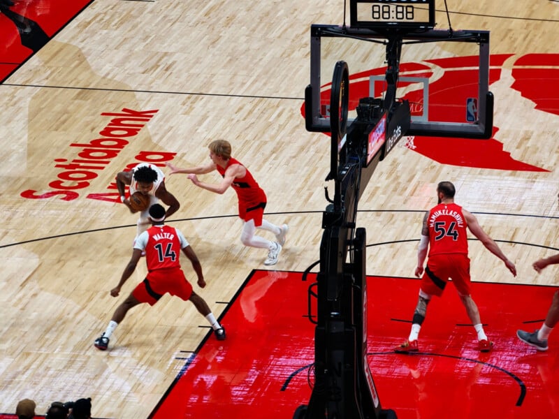 A basketball game at Scotiabank Arena shows a player in red dribbling while being closely guarded by an opposing player in white and red. Two other players in red jerseys are positioned nearby on the court.