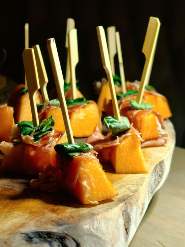 Several skewers with chunks of orange melon, thin prosciutto, and fresh basil are arranged on a rustic wooden board, creating an elegant appetizer display.