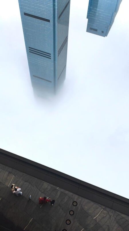 A tall skyscraper rises above thick white clouds, with only its top visible. Below, people stand on a dark rooftop at an angle, with another building partially visible in the mist.