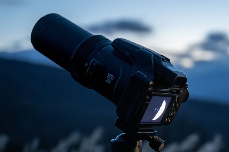Close-up of a camera on a tripod facing a moonlit sky. The digital display shows a crescent moon, while the background features a blurred landscape with soft dusk lighting.
