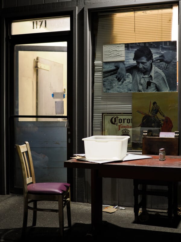A dimly lit storefront with a numbered door, a table, and a chair outside. There is a white container on the table and posters, including a vintage photo, on the window. A cardboard box and small bottle sit beside the container.