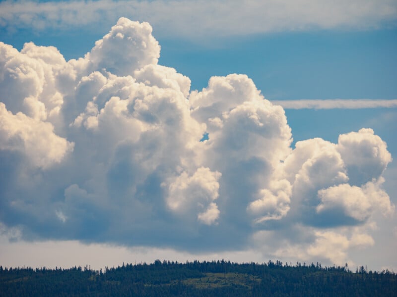 Large, fluffy white clouds rise dramatically into a blue sky above a forested hilltop, creating a vibrant and peaceful landscape.