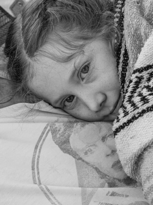 A young girl with light hair lies down, partially wrapped in a patterned blanket, looking directly at the camera. The image is in black and white. A graphic face print is visible on the shirt beneath her head.