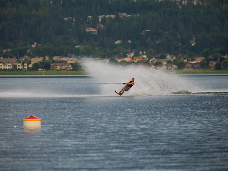 A person is water skiing on a lake, creating a large spray behind them. There are houses and trees on the far shore, and a yellow and red buoy floats in the foreground.