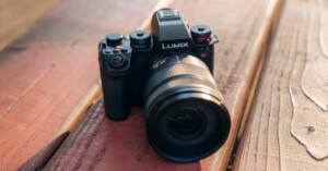 A black Panasonic Lumix S1 camera with a large lens sits on a wooden surface, angled to show the lens and control dials in natural light.