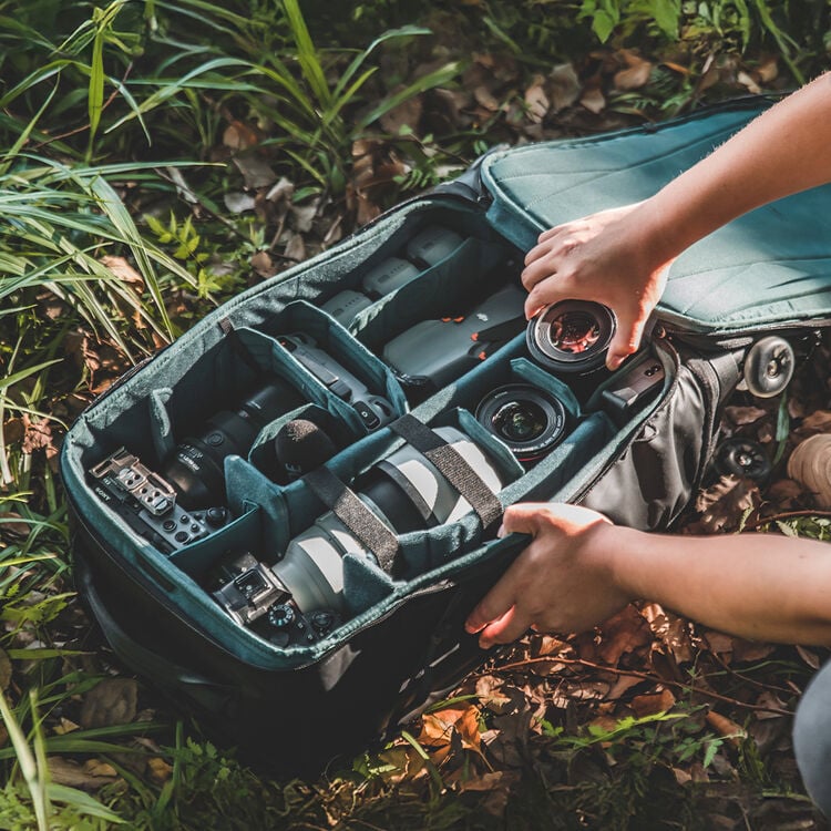 A person organizes camera equipment and lenses in a padded camera bag while outdoors, surrounded by grass and leaves.