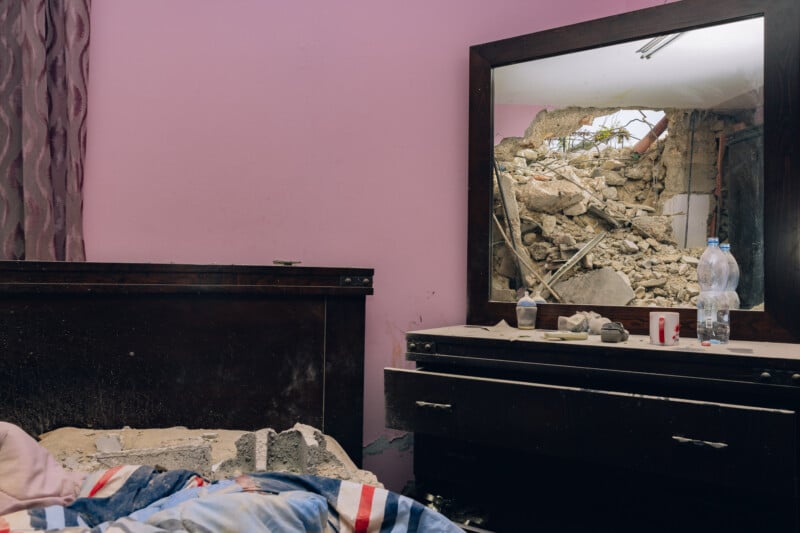 A bedroom with a pink wall and messy bed is shown. The mirror on the dresser reflects a collapsed wall and rubble, indicating severe damage outside the room. There are items scattered on the dresser.