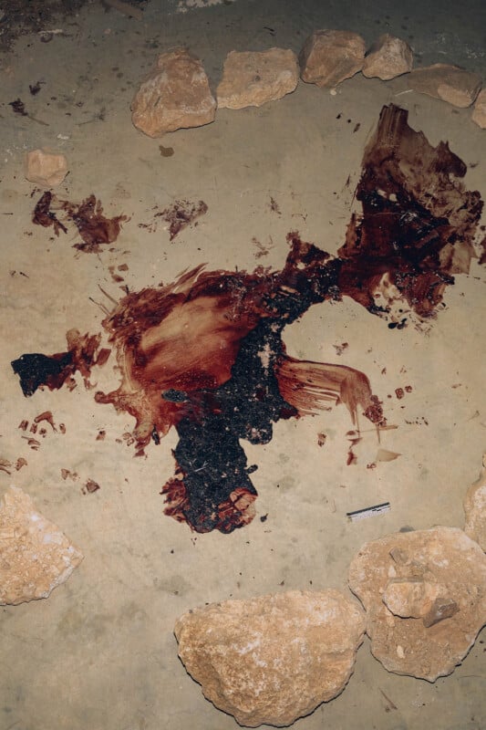 A patch of dark, dried blood and what appears to be charred material spreads across a concrete floor, surrounded by large, uneven rocks. A small ruler is placed near the stain for scale.