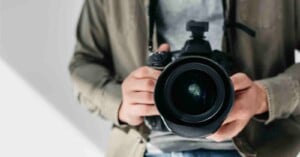 A person wearing a gray shirt and olive jacket holds a large professional camera, pointing the lens directly at the viewer. The background is neutral and out of focus.