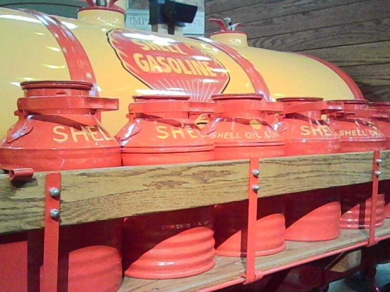 Row of vintage red and yellow Shell gasoline cans displayed on a wooden shelf in front of a large, matching Shell gasoline storage tank.
