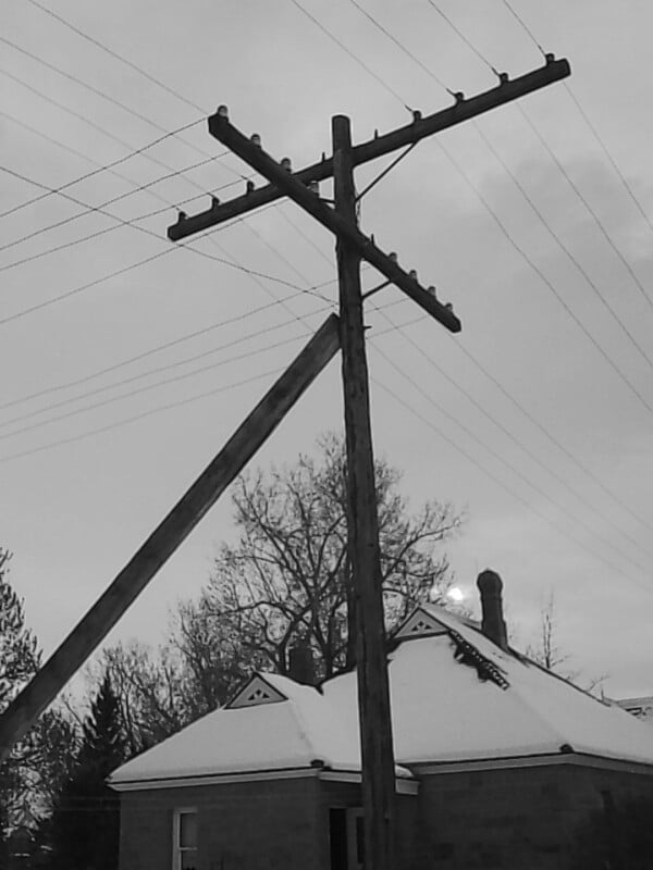 A black and white photo of a utility pole with wires, positioned in front of a house with a snowy roof. Leafless tree branches and a mostly cloudy sky appear in the background.