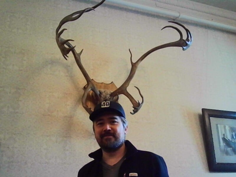 A man wearing a black jacket and cap stands in front of a wall with large mounted antlers, making it appear as if the antlers are growing from his head. There is also a framed picture on the wall to the right.
