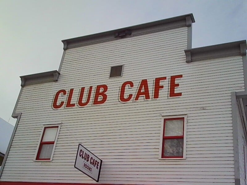 A white building with red-trimmed windows features large red letters spelling "CLUB CAFE" on the front. A slightly tilted sign below also reads "CLUB CAFE" with a smaller word "ROOMS" underneath.