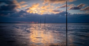 Sunset over a wet, reflective beach with dramatic clouds. Several wooden poles in a line extend into the distance, creating depth and leading the eye towards the horizon. The sky glows with orange and blue hues.