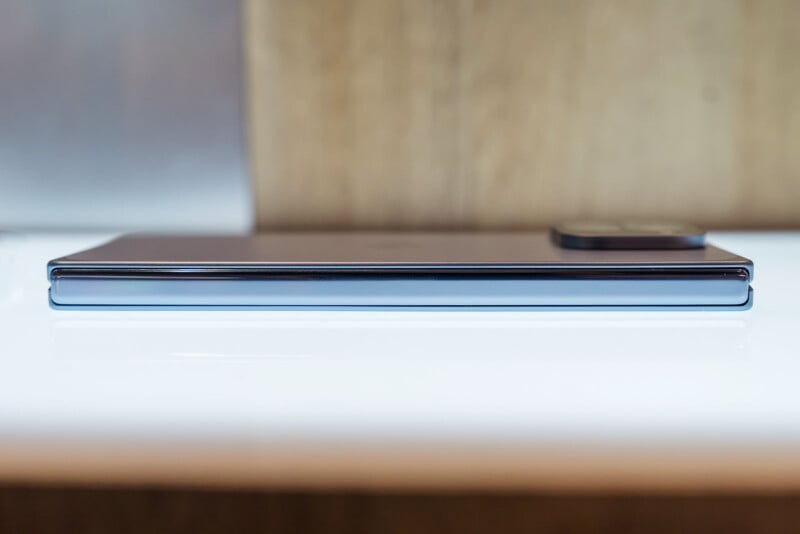 A modern smartphone lies flat on a white surface, viewed from the side, highlighting its slim profile and protruding rear camera module, with a blurred brown background.