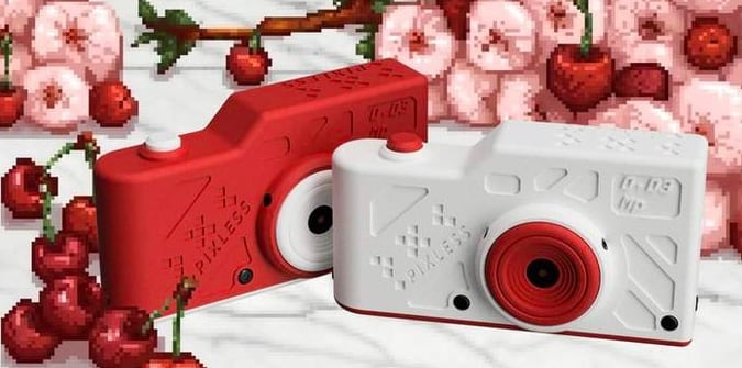 Two toy cameras, one red and one white with red accents, are placed in front of a background featuring pixel art cherries, strawberries, and pink clouds. The cameras have a playful, blocky design.