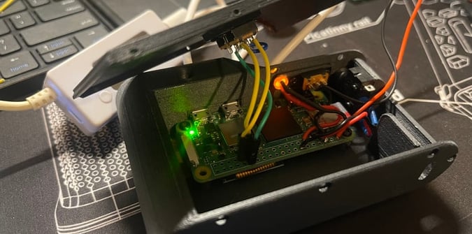 A small electronic device with a green circuit board, various colored wires, and indicator lights sits inside a partially open black plastic case near a keyboard.