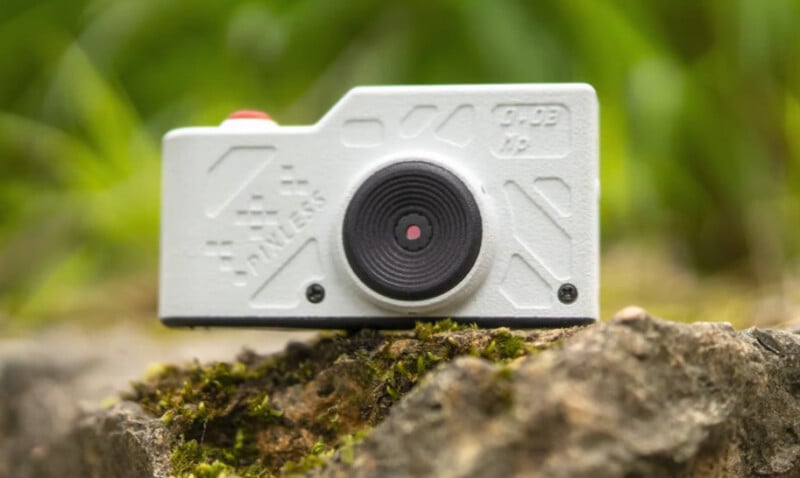 A small white camera with a textured surface and a large lens, labeled "RUVLens," sits on a mossy rock with green foliage blurred in the background.