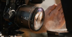 Close-up of a camera with a large 42mm lens in focus, capturing a scene. A hand is visible in the background, partially blurred, with sunlight reflecting off the lens and a bit of smoke or mist rising nearby.