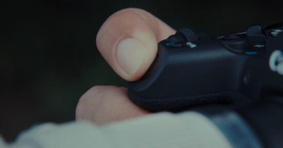 A close-up of a person's hand holding a black game controller, with their thumb resting on one of the buttons.