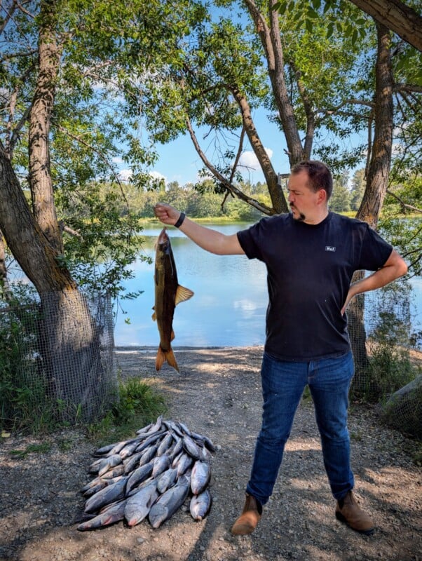 A man in a black shirt and jeans stands by a lake, holding up a large fish with one hand. At his feet, there is a pile of caught fish. Trees and water are visible in the background.