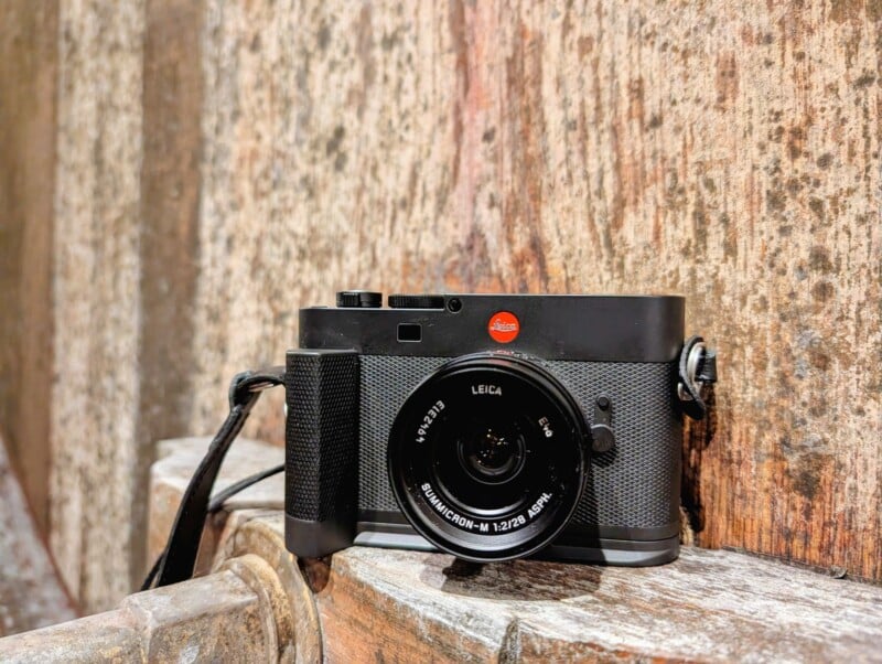 A black Leica camera with a lens attached rests on a wooden surface against a textured, rustic wooden background.