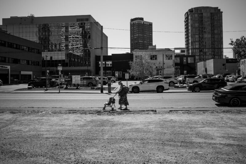 A woman and a child walk across a street in an urban area, with tall buildings, parked cars, and reflections in the glass windows in the background. The image is in black and white.