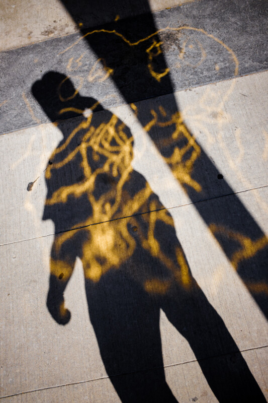 A person's shadow is cast on a sidewalk covered with bright yellow spray-painted markings and lines, with contrasting light and dark areas from surrounding structures.